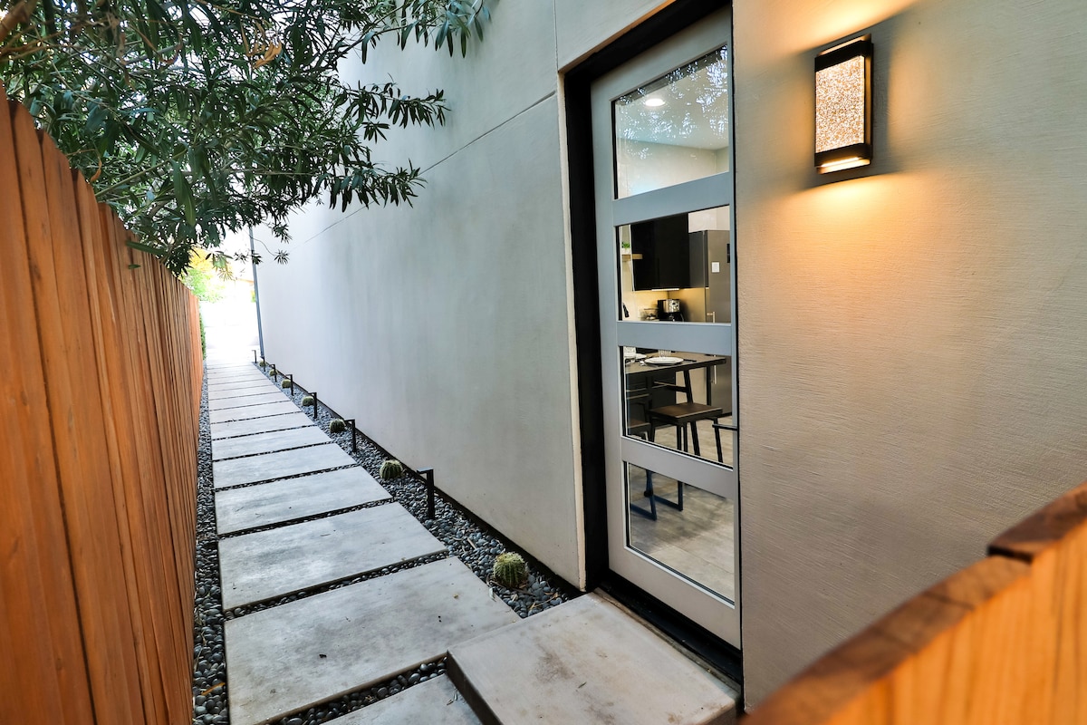 The entrance is framed by a wooden fence and features a paved walkway leading to a glass door. Soft lighting from a wall sconce highlights the sleek modern design, while small decorative stones and plants line the pathway, creating a welcoming approach.