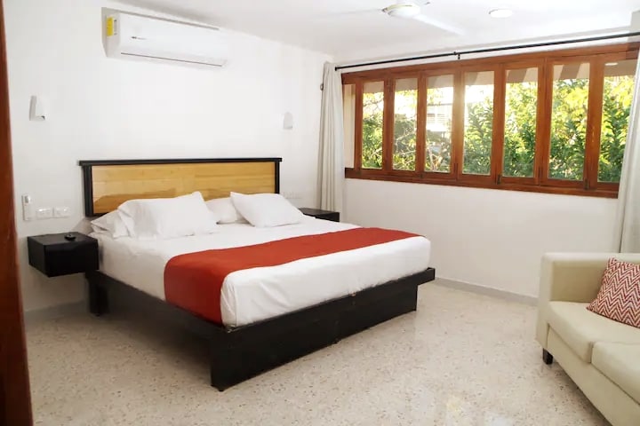 A spacious sleeping area is highlighted by a large bed dressed in crisp white linens and a vibrant red accent blanket. Multiple windows allow natural light to fill the room, with greenery visible outside. A cozy seating area is present, featuring a light-colored sofa with decorative cushions.