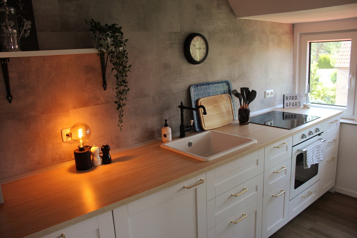 The kitchen features modern cabinets with a light wood countertop. A sink is positioned under a window, allowing natural light. Various kitchen tools are organized neatly, with decorative plants and a clock adding subtle accents. A dishwasher and oven are present, completing the functional space.