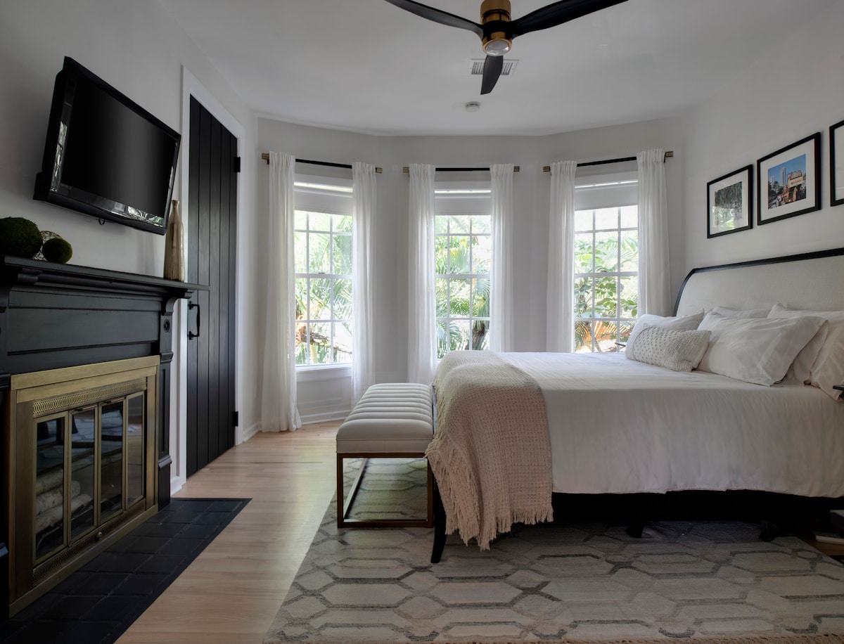 A spacious bedroom features a king-sized bed with soft bedding. Natural light filters through large bay windows, adorned with sheer white curtains. A cozy ottoman is positioned at the foot of the bed, complemented by a decorative fireplace and a wall-mounted TV.