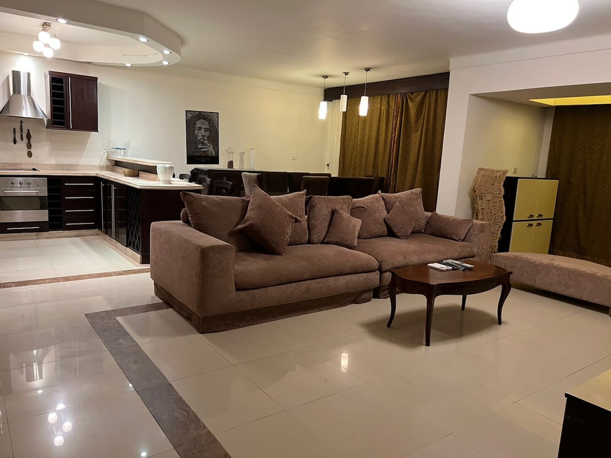 A spacious living area showcases a large, plush sectional sofa with decorative cushions positioned around a dark wooden coffee table. An open kitchen with modern appliances is visible in the background, while warm lighting creates a cozy ambiance throughout the room.