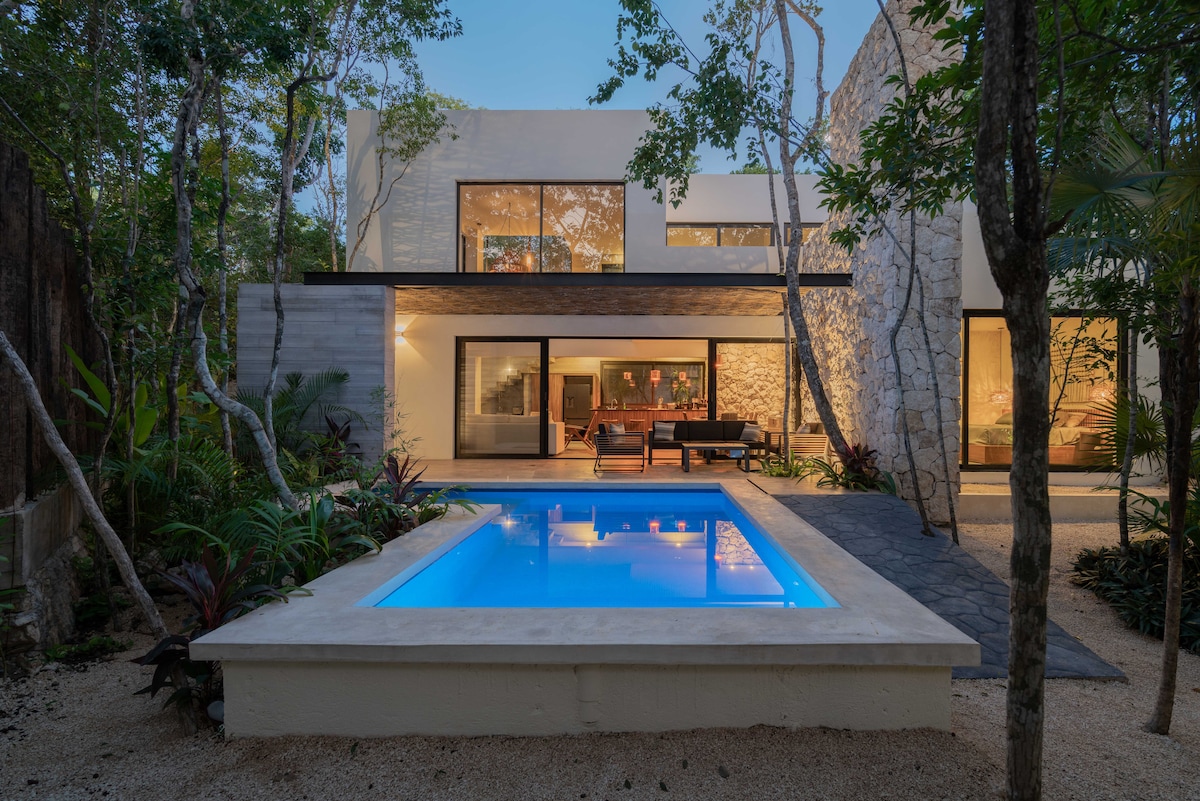 A private pool is positioned in front of a modern two-story home, surrounded by lush greenery. Large glass doors invite natural light into the living spaces, while a comfortable lounge area is visible on the patio, creating a serene ambiance within the jungle setting.