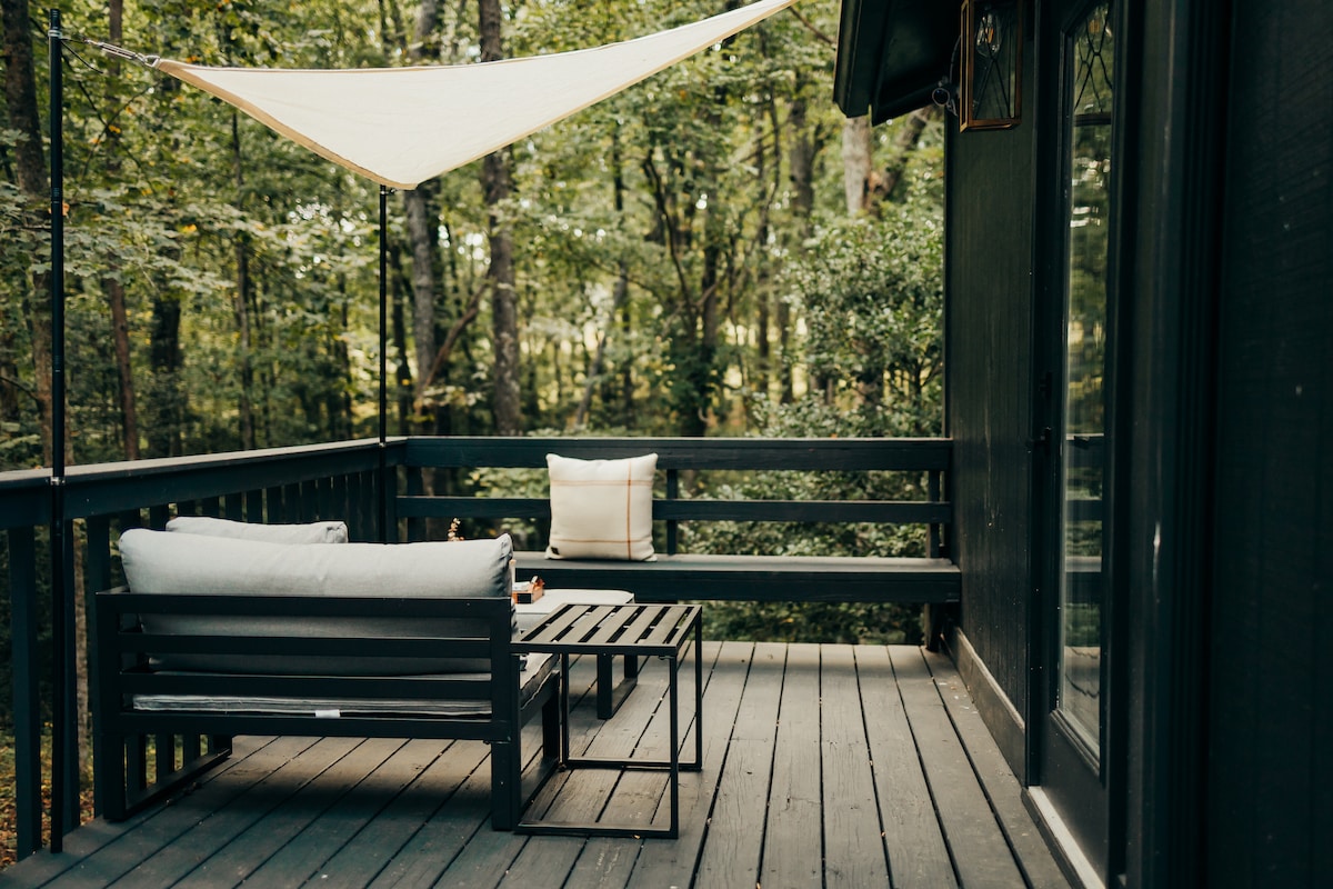 A side deck area features a simple seating arrangement, including a low sofa with cushions and a coffee table. Shade is provided by a sail awning, while surrounding trees create a natural, wooded backdrop.