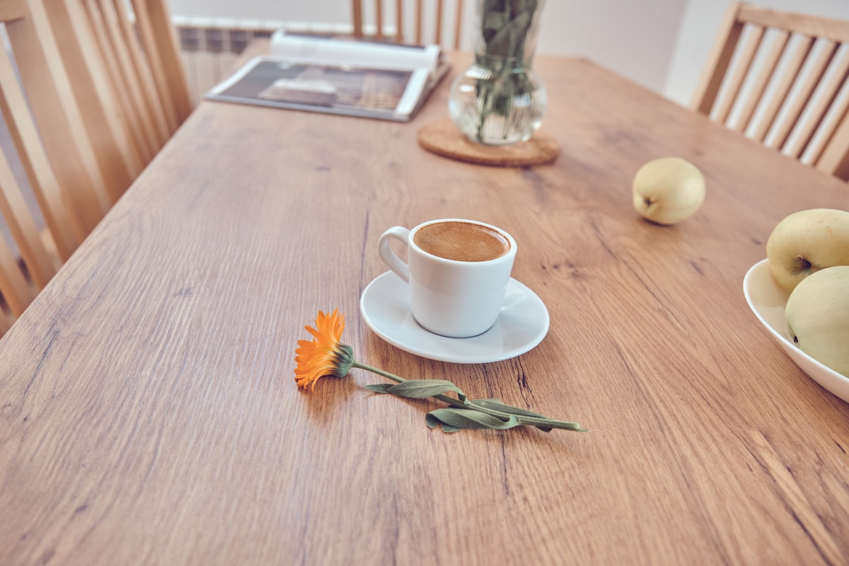 A wooden dining table features a small white cup filled with coffee on a matching saucer. A vibrant orange flower rests beside the cup, while a bowl of apples sits in the background. A blurred magazine and a vase with greenery are visible on the table.