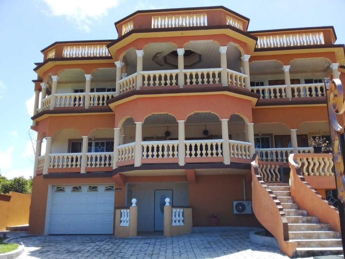 The exterior of a stylish multi-level building features decorative balconies with white railings. An entrance with a staircase leads to the door, complemented by a garage and an air conditioning unit adjacent to the main structure, all under a clear blue sky.