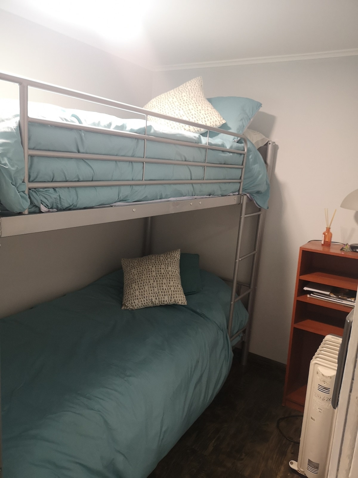 A compact sleeping area is equipped with a bunk bed featuring two comfortable mattresses dressed in soft blue bedding. A decorative pillow is placed on each bed. A small bedside table and lamp are positioned next to the bottom bed, complementing the minimalist decor.