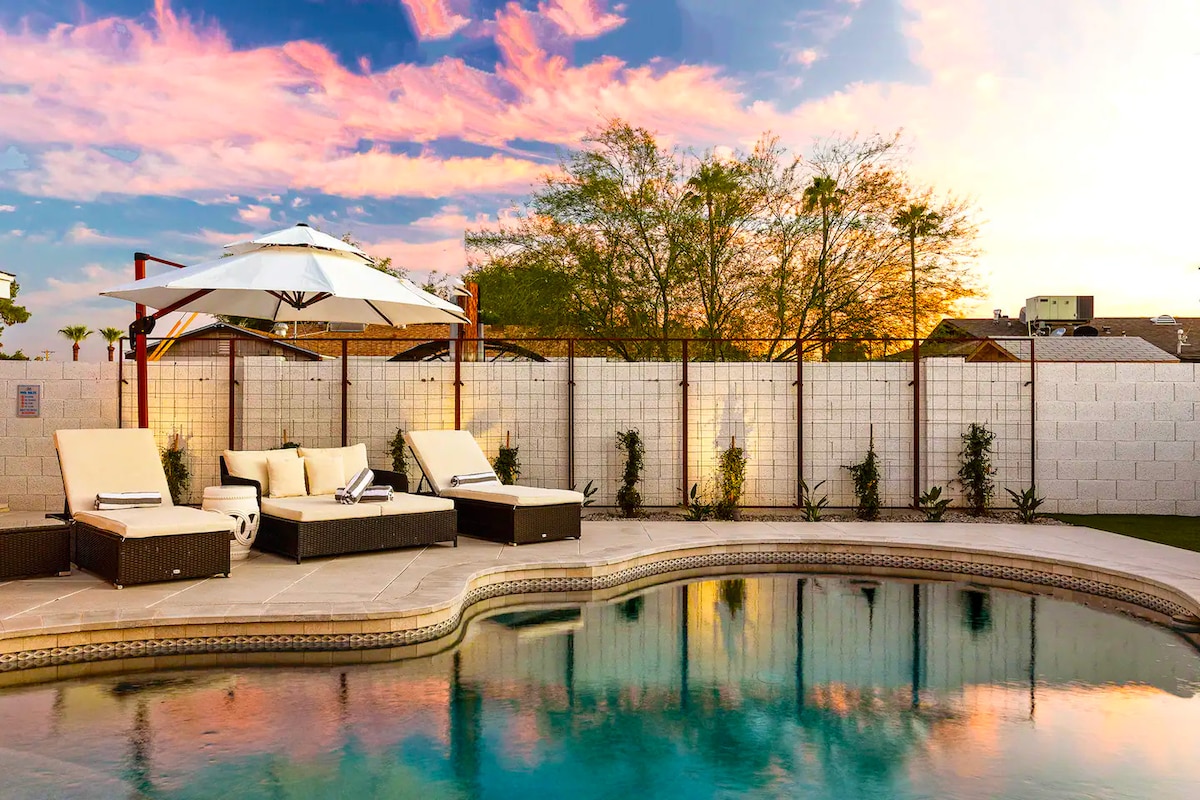 A heated pool is surrounded by modern lounge chairs and a large umbrella. The water reflects the colorful sky at dusk, while the landscaped area is complemented by several green plants along the perimeter.