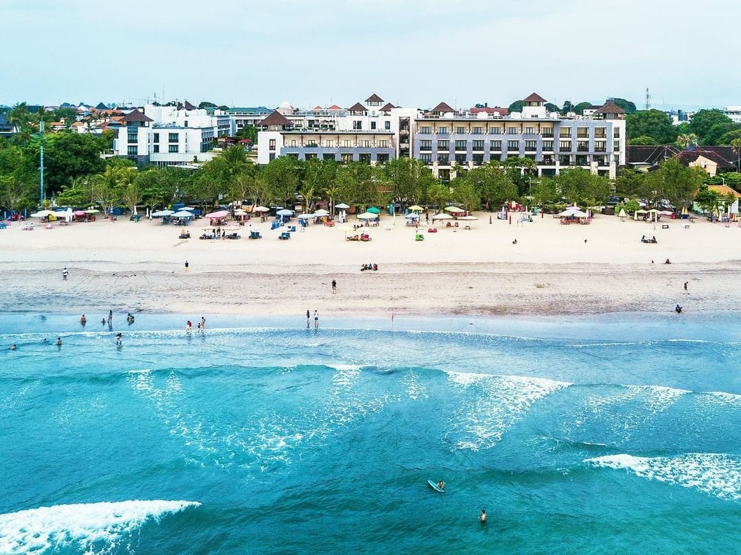 The beachfront property is positioned along Kuta Beach, surrounded by smooth sands and the gentle waves of the ocean. Sunbathers and beachgoers enjoy the outdoor space, while the hotel's multiple levels and landscaped areas provide a scenic backdrop.