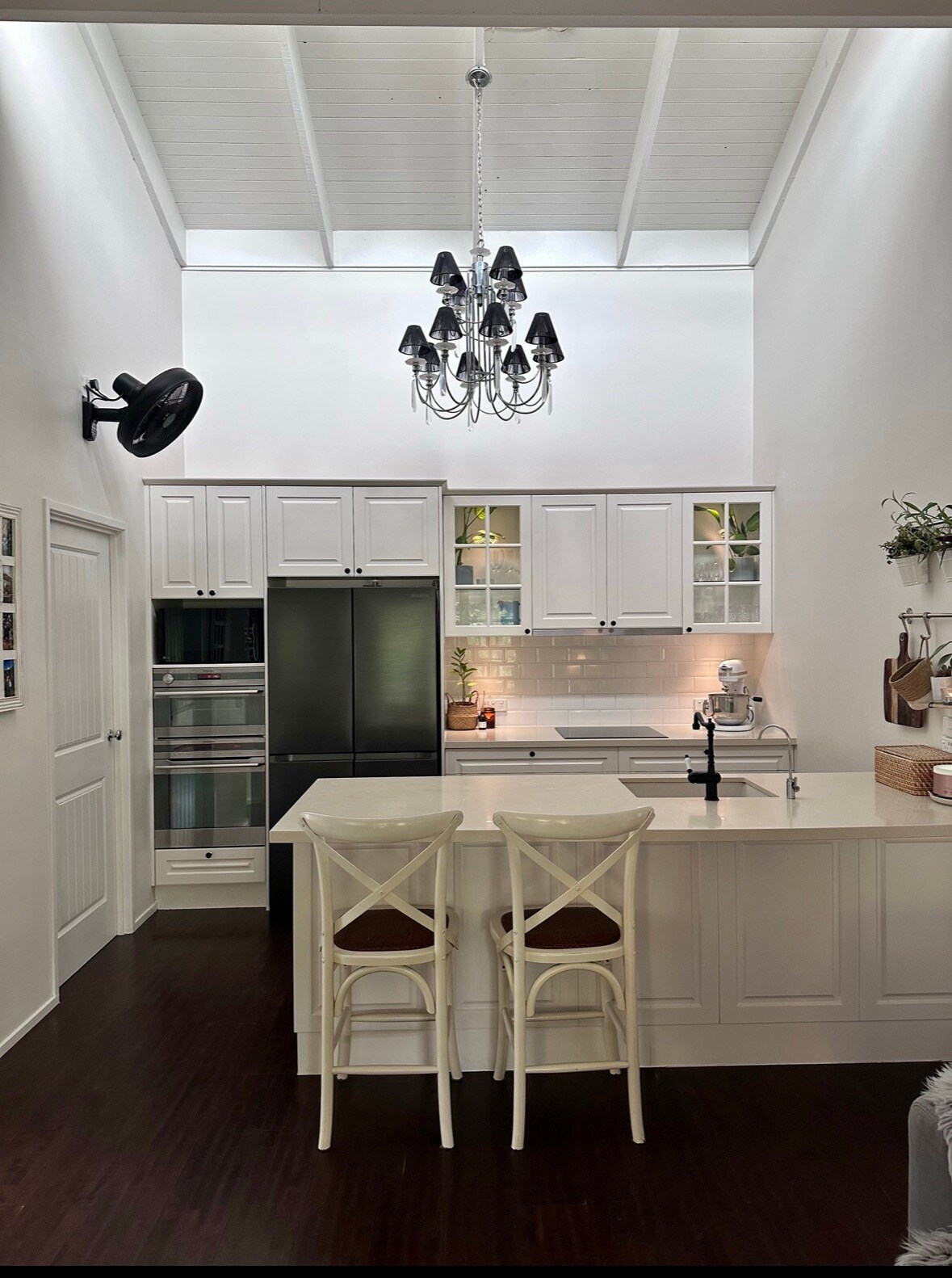 A modern kitchen is highlighted by tall white cabinetry and a central island with two stools. A sleek black refrigerator stands beside a built-in oven, while plants decoratively accent the shelves. A chandelier adds elegance to the space, with natural light enhancing the inviting atmosphere.