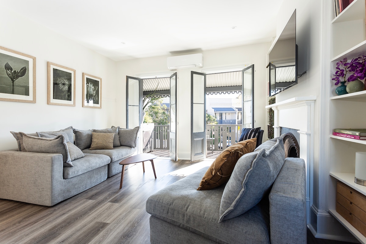 The open plan living area features a spacious grey sectional sofa and a wooden coffee table. Large French doors open to a sunlit balcony, enhancing the natural light. Simple artwork adorns the walls, adding a touch of sophistication to the airy space.