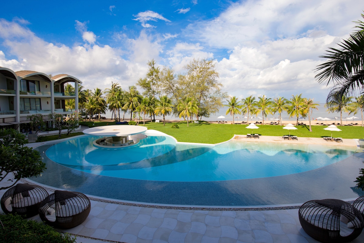 A serene outdoor area features a curved swimming pool, blending seamlessly with the surrounding lush green grass. Palm trees provide natural shade, and several lounge chairs are arranged under umbrellas, offering inviting spots for relaxation. A clear sky is visible, complementing the tranquil setting.