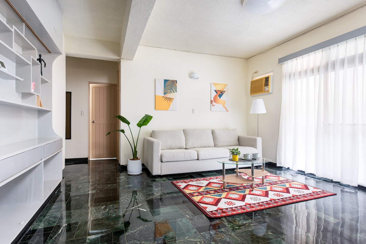 A bright living room is featured, with a comfortable sofa facing a glass coffee table. Decorative artwork adorns the walls, and a potted plant adds a touch of greenery. Natural light filters through sheer curtains, illuminating the glossy tiled floor.