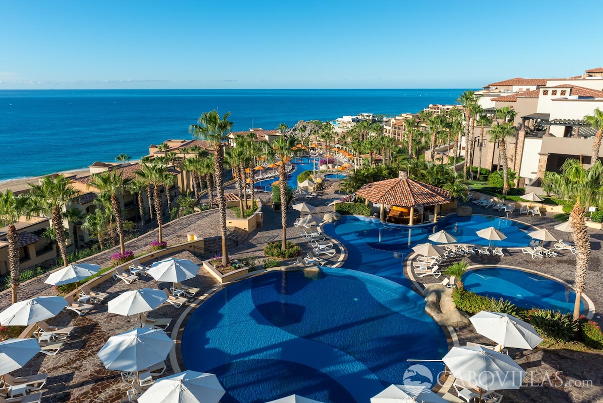 A wide view showcases the resort's various pools surrounded by palm trees and sun loungers. Umbrellas provide shade for guests while the vast ocean is visible in the background, under a clear sky. The inviting scenery emphasizes the relaxation potential of the resort.