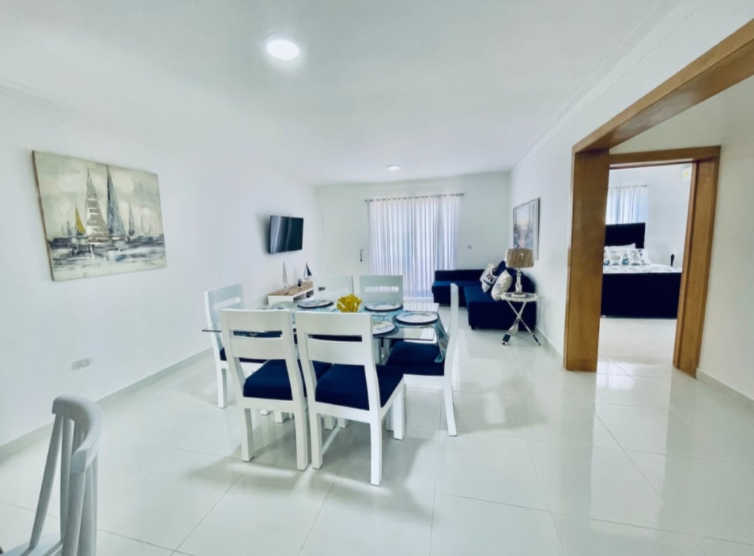 A bright and airy living area features a glass dining table surrounded by white chairs, complemented by a modern sofa. Light-colored walls enhance the space, and a large window allows natural light to fill the room. An art piece adorns one wall, adding visual interest.
