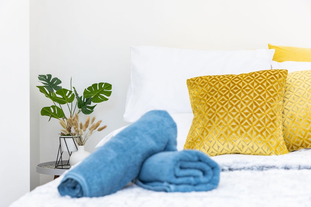 A neatly arranged bed features plush white pillows and vibrant throw pillows in yellow and geometric patterns. Soft rolled towels in shades of blue are placed nearby, while a small plant adds a touch of greenery on a side table, complementing the bright and fresh atmosphere.