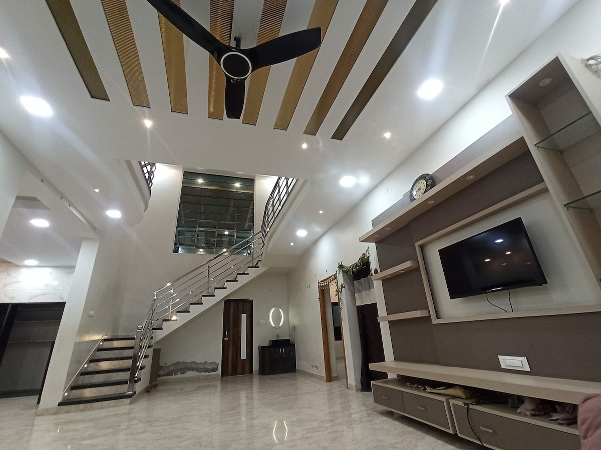 A spacious living area features a modern design with high ceilings and sleek lighting. A stylish staircase leads to an upper level, while a wall-mounted television is positioned above elegant cabinetry. Large windows allow natural light to fill the space.