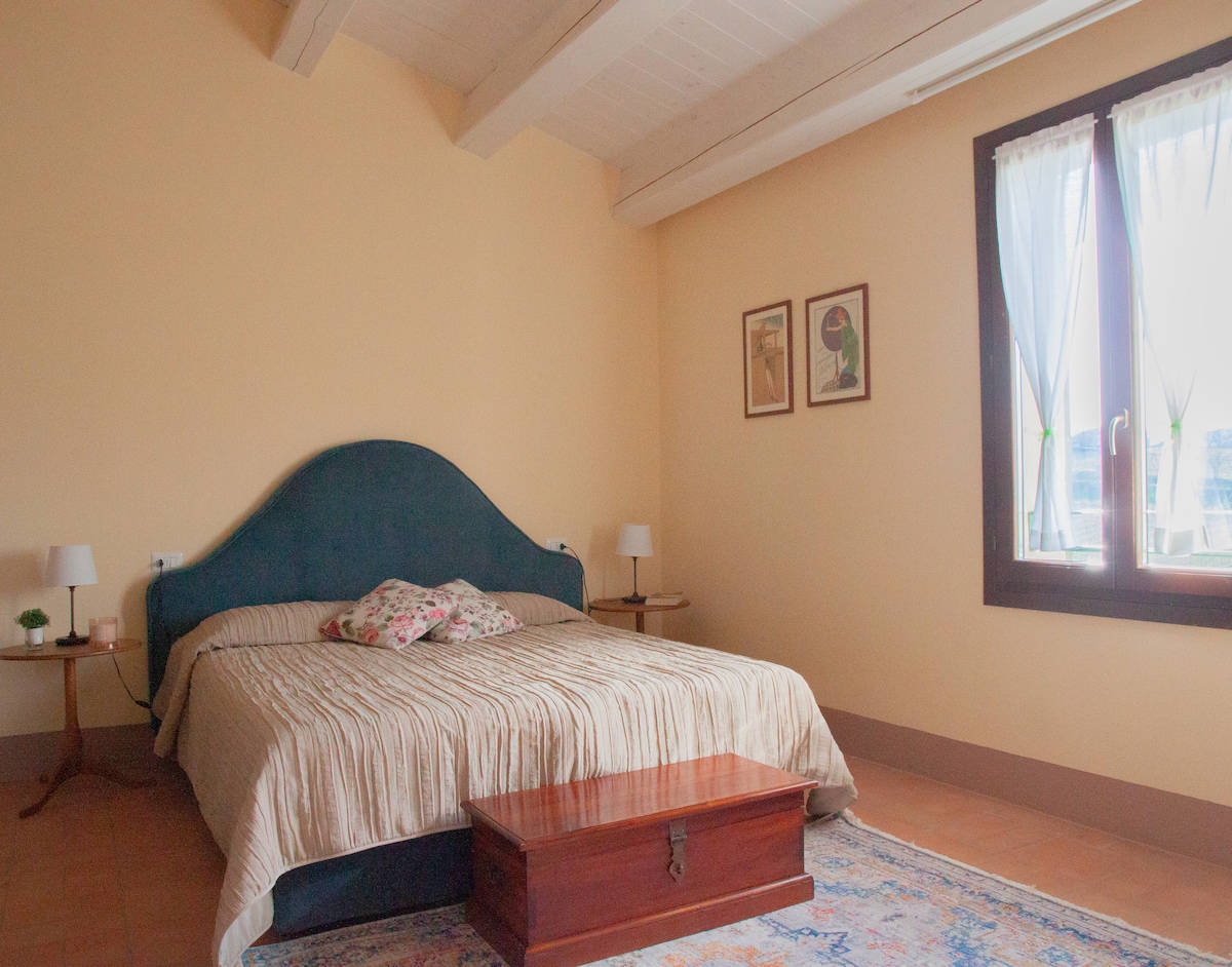 A warm-toned bedroom features a spacious bed with a light-colored quilt and decorative pillows. Two bedside tables with lamps sit beside the bed, while a wooden coffee table is positioned in front. Natural light filters through a pair of windows adorned with sheer curtains.