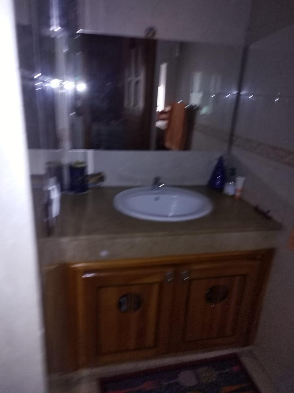 A bathroom sink area is showcased, featuring a circular basin set in a stone countertop with wooden cabinetry below. A mirror reflects a glimpse of the room beyond, while toiletries and personal items are neatly arranged beside the sink.
