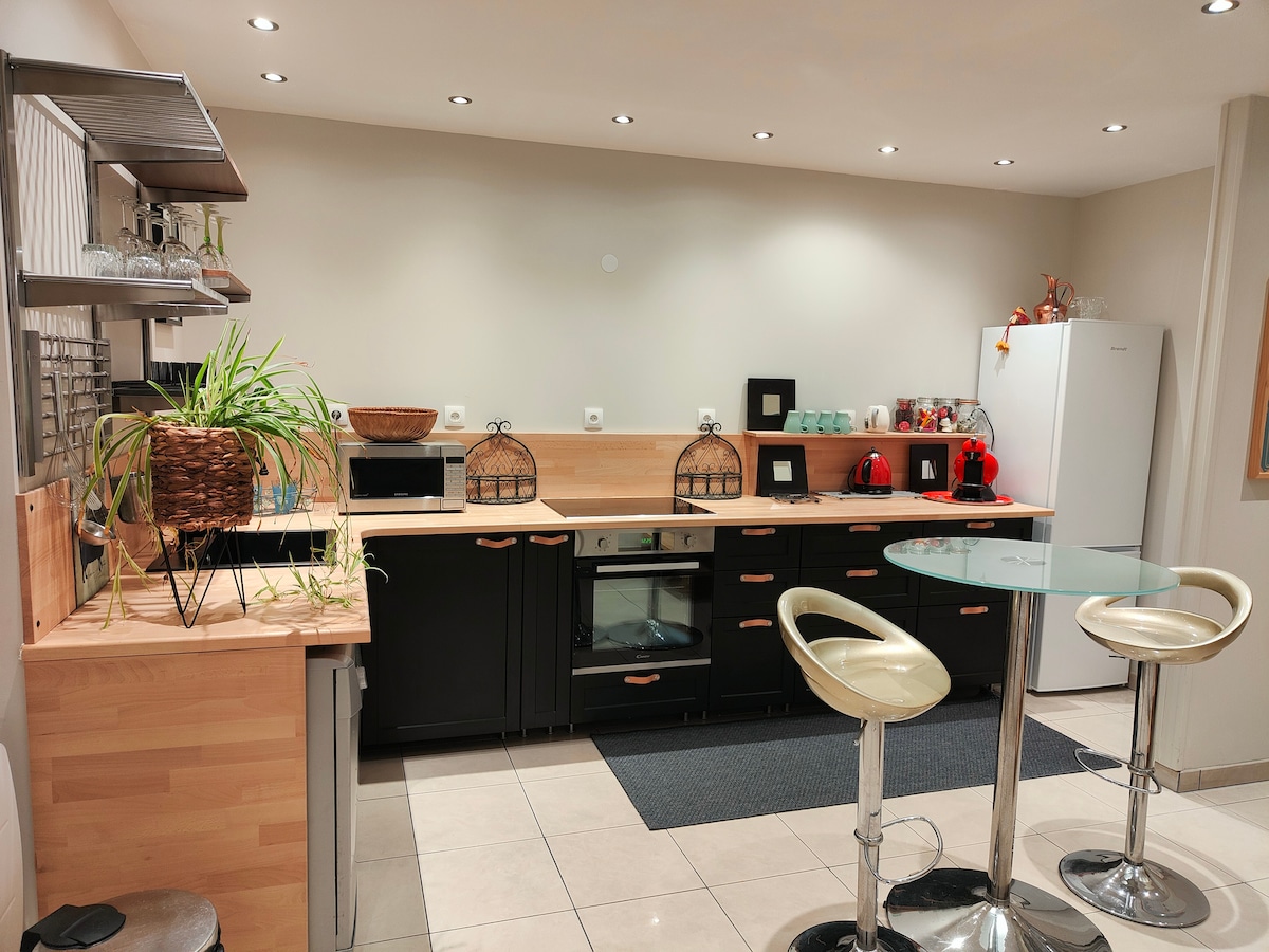 The kitchen features modern cabinetry in a dark finish with wooden accents. A sleek countertop holds a microwave and an oven, while a refrigerator stands against the wall. Bar stools are situated at a circular glass table, and potted plants add a touch of greenery.
