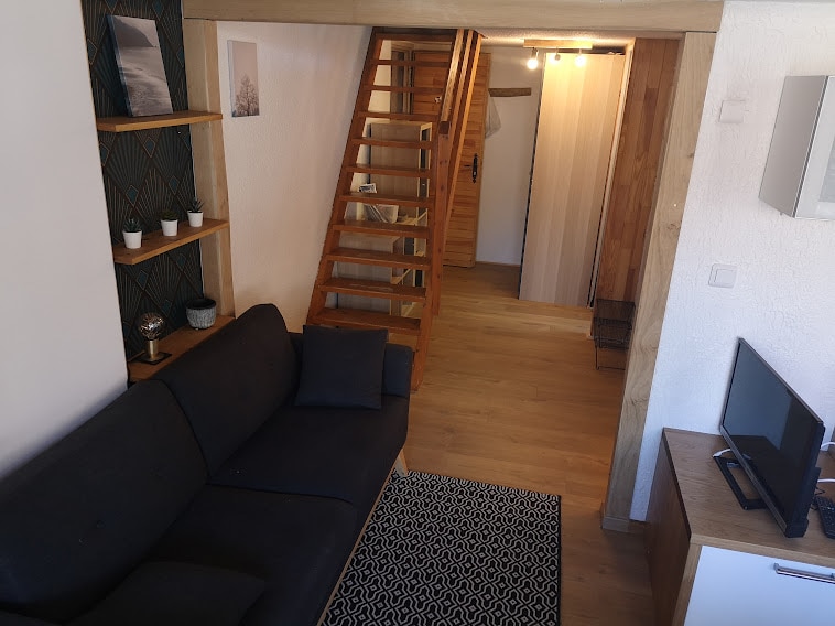 A wooden staircase leads to an upper level, framed by light-colored walls. A dark sofa is positioned on the left, complemented by a decorative rug. A TV is placed on a light wooden cabinet across from the sofa, contributing to a cozy living space.