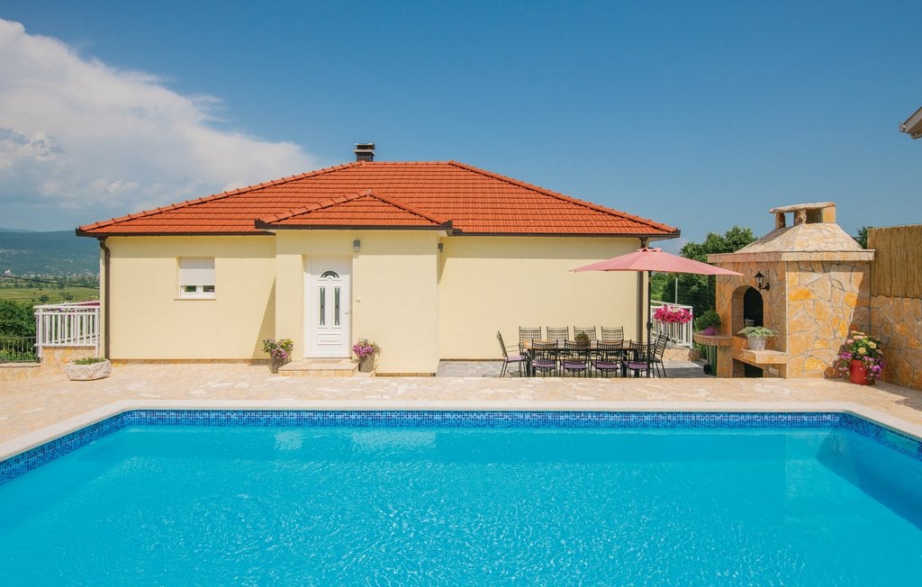 A charming villa with a red-tiled roof is situated beside a clear blue pool. Outdoor seating is arranged around a dining table under an umbrella, and a stone grill is nearby, providing an inviting space for gatherings. Lush greenery is visible in the background.