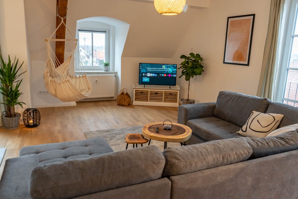 A spacious living area is illuminated by natural light from large windows. A comfortable sectional sofa is paired with a stylish coffee table. A hanging chair adds a cozy touch, while indoor plants contribute to a refreshing ambiance. A large television is ready for entertainment.