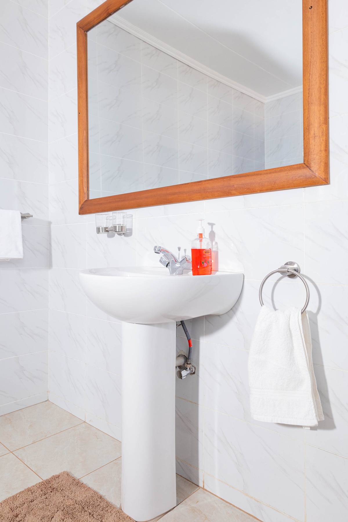 A clean and well-lit bathroom is displayed, featuring a white sink with a modern faucet. A framed mirror hangs above the sink, reflecting the space. A white towel is neatly hung on a towel rack, and a small rug lies on the tiled floor.