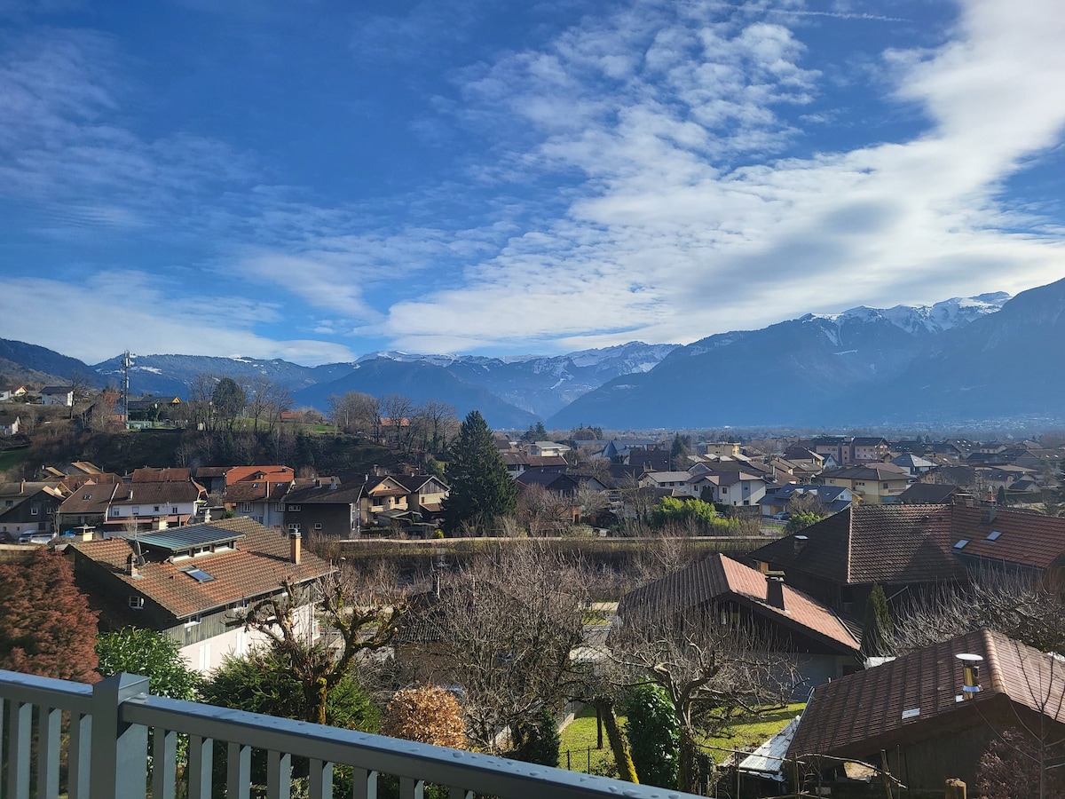 The view features a scenic landscape of rooftops nestled in a valley, surrounded by rolling hills and majestic mountains. A clear blue sky is interspersed with soft clouds, creating a serene backdrop for the charming village below.
