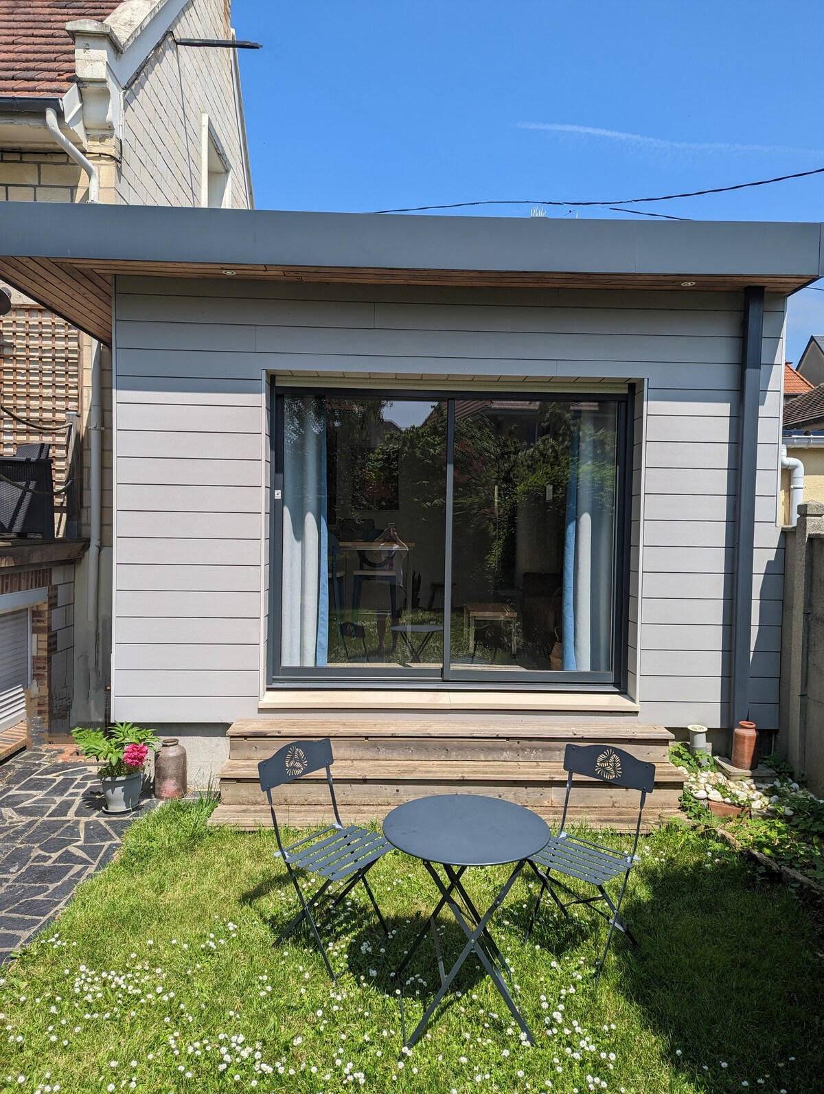 A modern exterior of a small house is depicted, featuring a large glass window framed by light-colored siding. A small outdoor seating area is visible, consisting of a round table and two chairs set on a grassy patch, surrounded by blooming flowers.