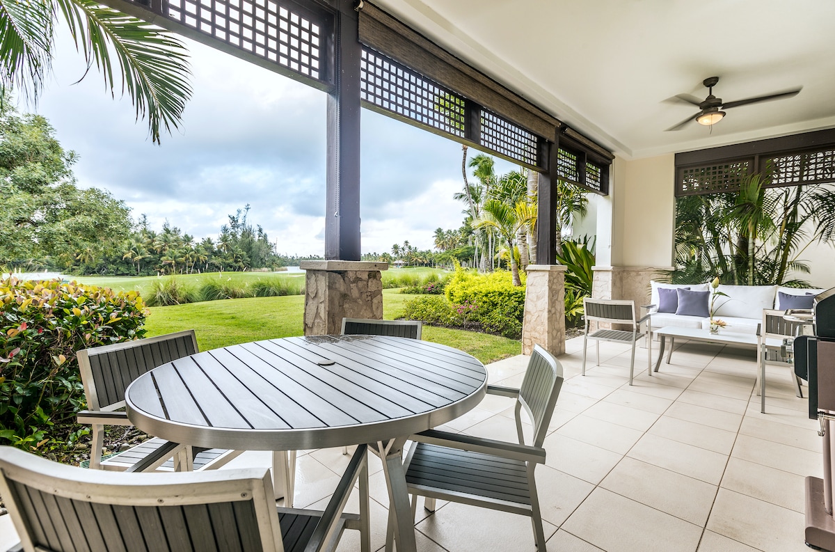 An outdoor terrace features a round table surrounded by six chairs, complemented by a seating area with two armchairs and a couch. Lush greenery and a golf course view can be seen through the open space, enhancing the tranquil ambiance of the setting.