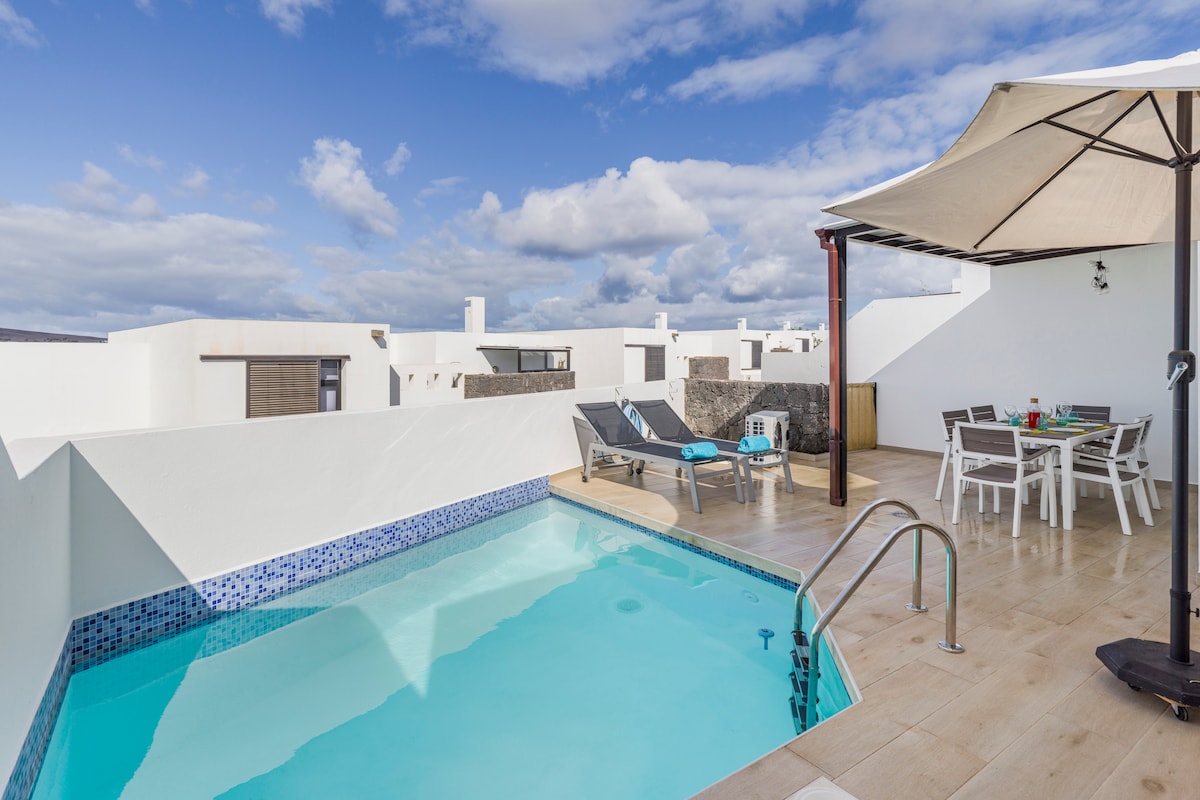 A modern terrace features a private swimming pool with clear, inviting water. Comfortable loungers are positioned nearby, with a shaded dining area set for meals. The surrounding white walls create a serene atmosphere, and the sky is adorned with soft clouds.