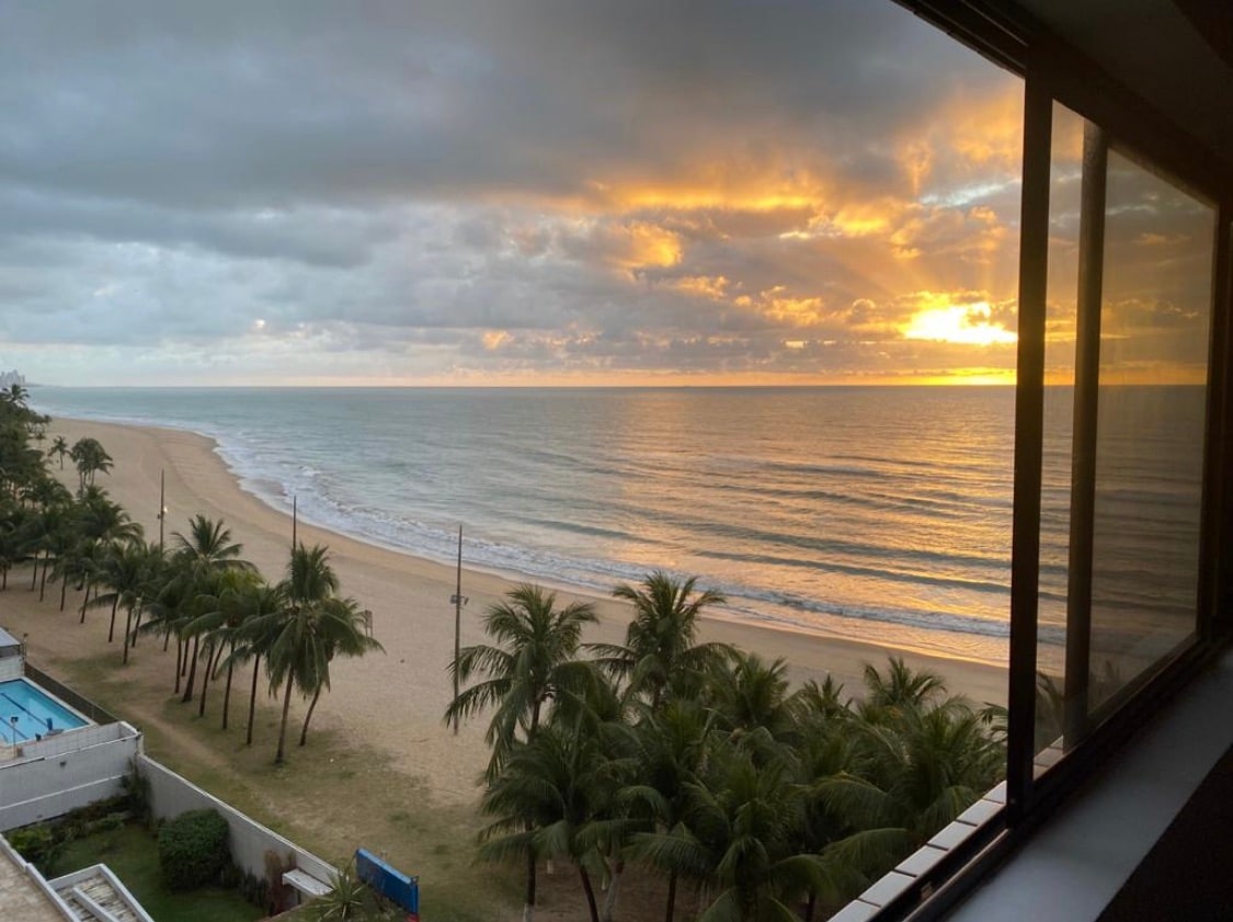 A sweeping view of the beach is captured from the window, with golden sunrise hues reflecting on the calm water. Palm trees line the shore, and the sandy beach extends along the horizon, creating a peaceful coastal scene.