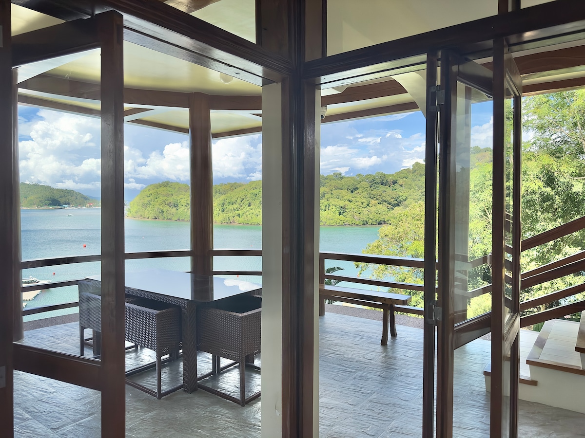 A covered terrace offers a seating area with woven chairs, surrounded by large glass doors. Views of the serene water and lush green hills are framed by the open space, allowing for a seamless connection to the outdoor environment.