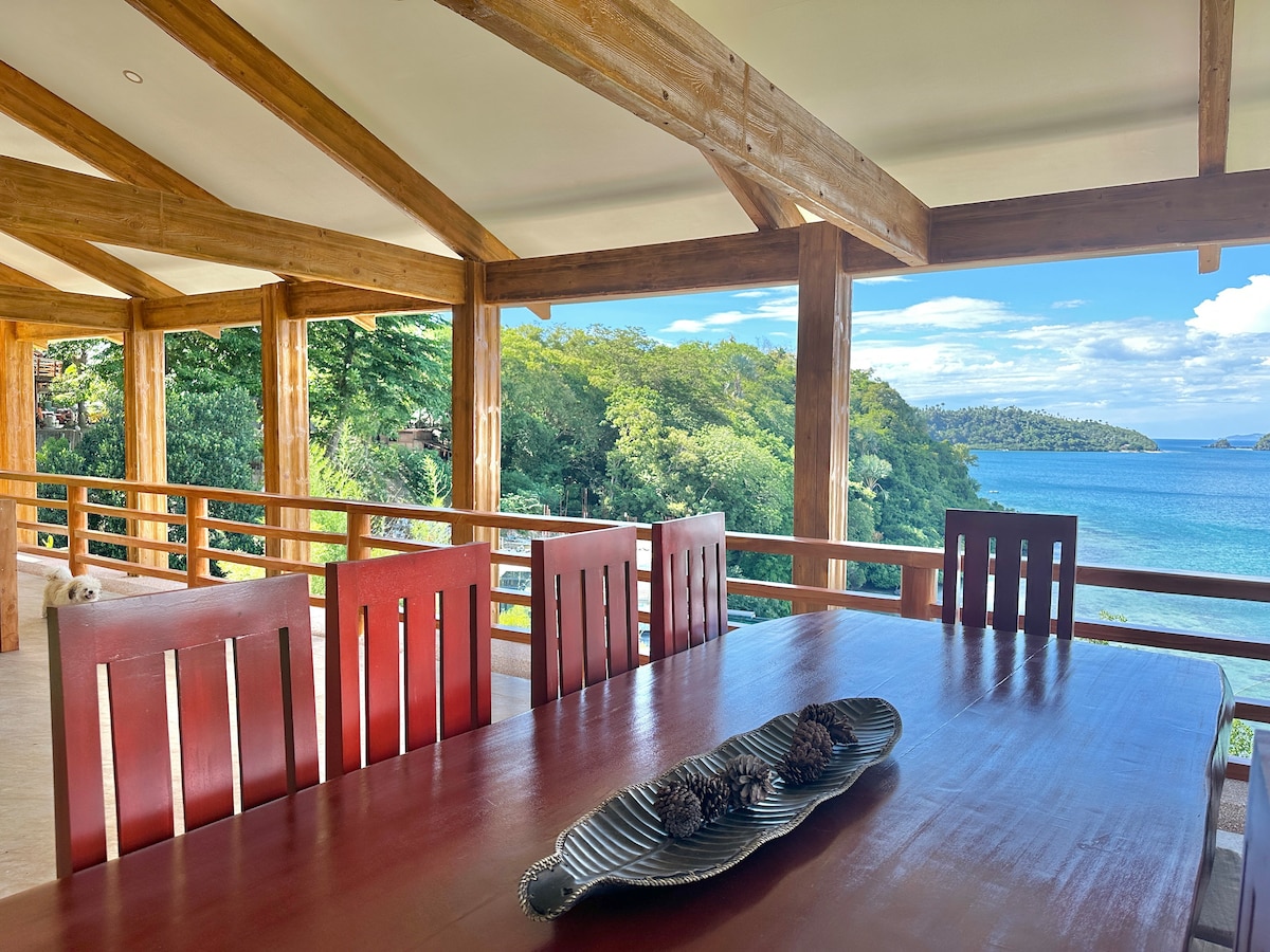 A large wooden dining table with six red chairs is positioned on a terrace. Expansive views of lush greenery and the blue waters of Puerto Galera Bay are visible through the open space, with beams of wood framing the scene.