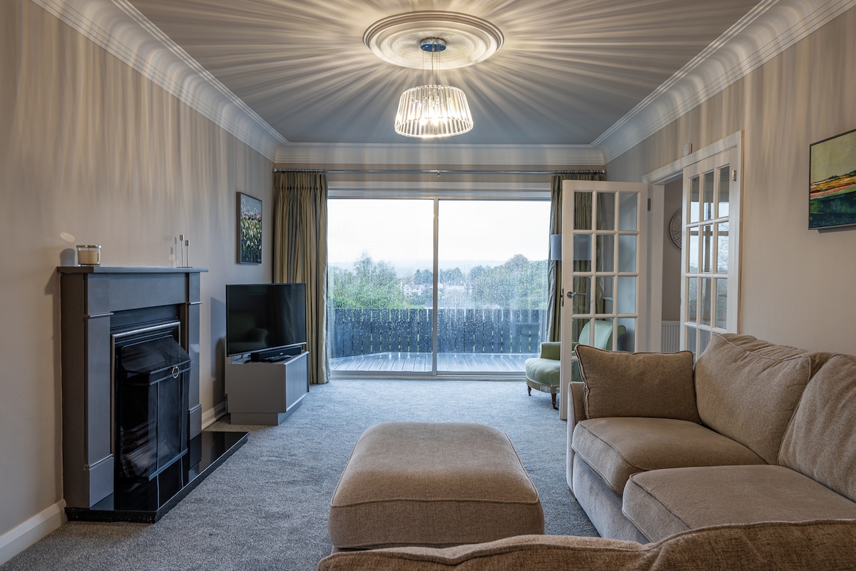 A spacious living room features a large sectional sofa and a cozy ottoman. A flat-screen television is positioned near a classic fireplace. Natural light pours in through expansive sliding doors, leading to a deck area. Tasteful wall decor adds a subtle touch to the neutral color scheme.