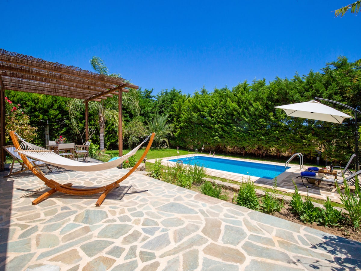 A serene outdoor space showcases a private pool surrounded by lush greenery. A hammock is gently swaying under a wooden pergola, while a sun-lounger sits beneath a large umbrella. A stone patio complements the inviting garden atmosphere.