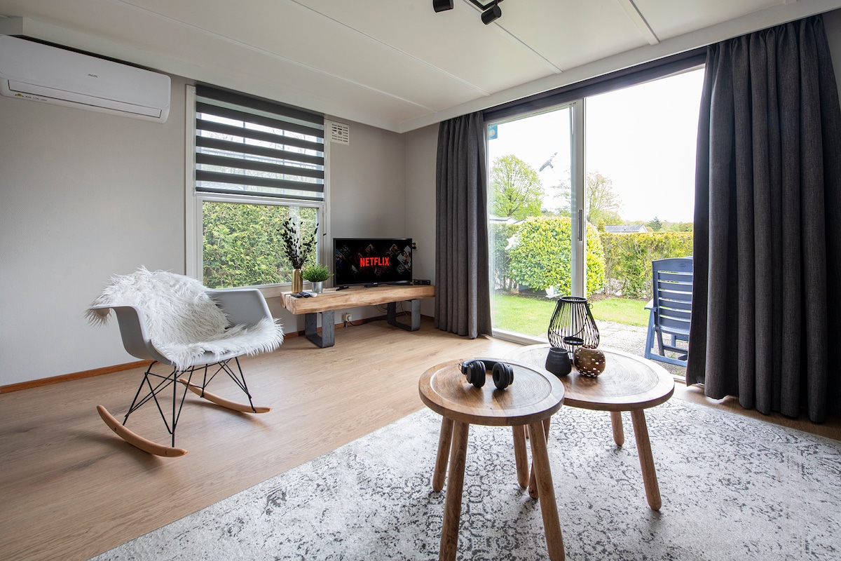 A bright living area features a rocking chair and two wooden tables. A television displays Netflix, and large glass doors open to the garden. Dark curtains frame the windows, while a cozy rug adds comfort to the space.
