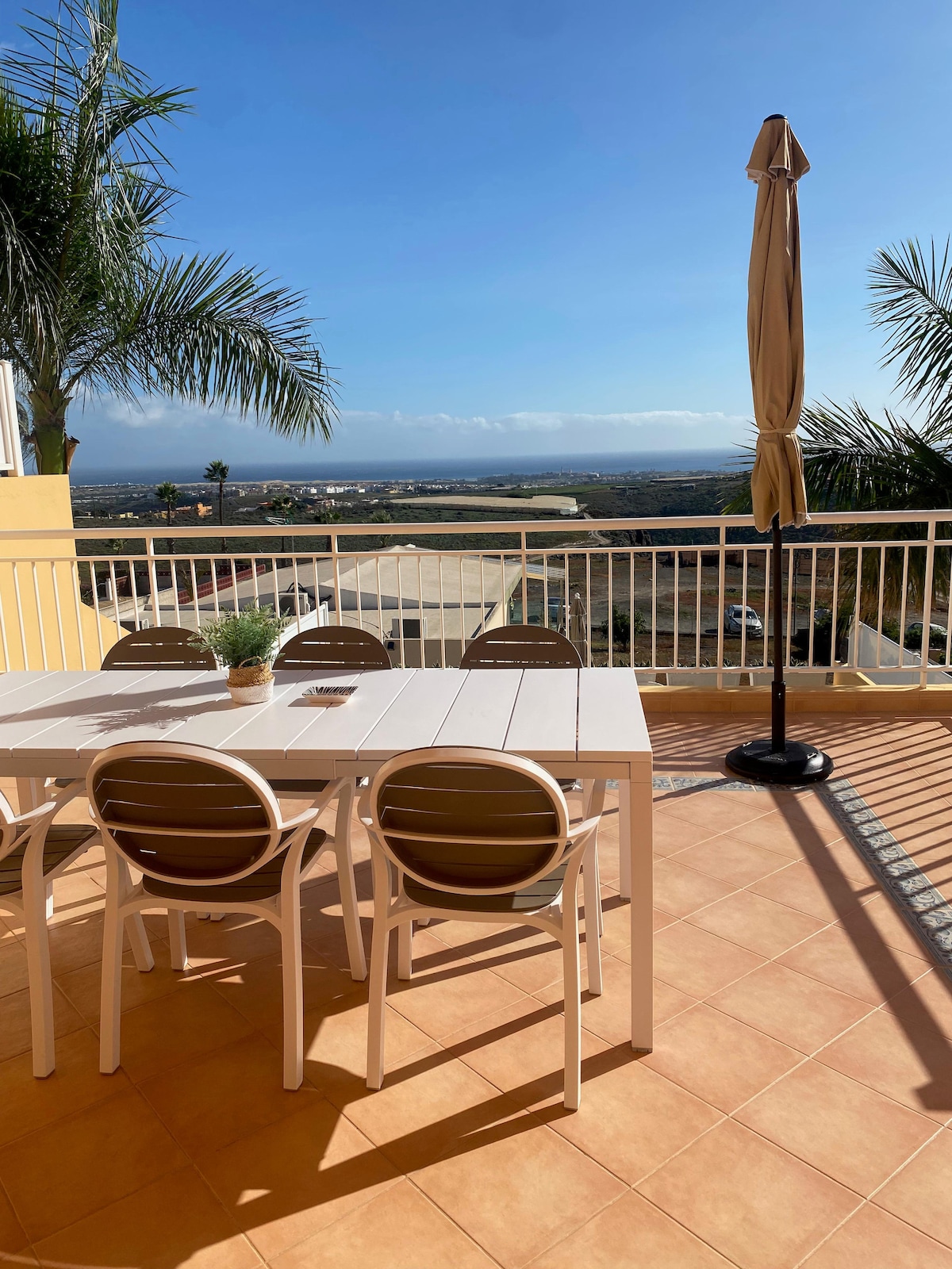 A spacious terrace features a large white dining table surrounded by six light-colored chairs. A sun umbrella stands nearby, providing shade. Lush palm trees are visible on the sides, and a panoramic view of the landscape stretches into the distance under a clear blue sky.