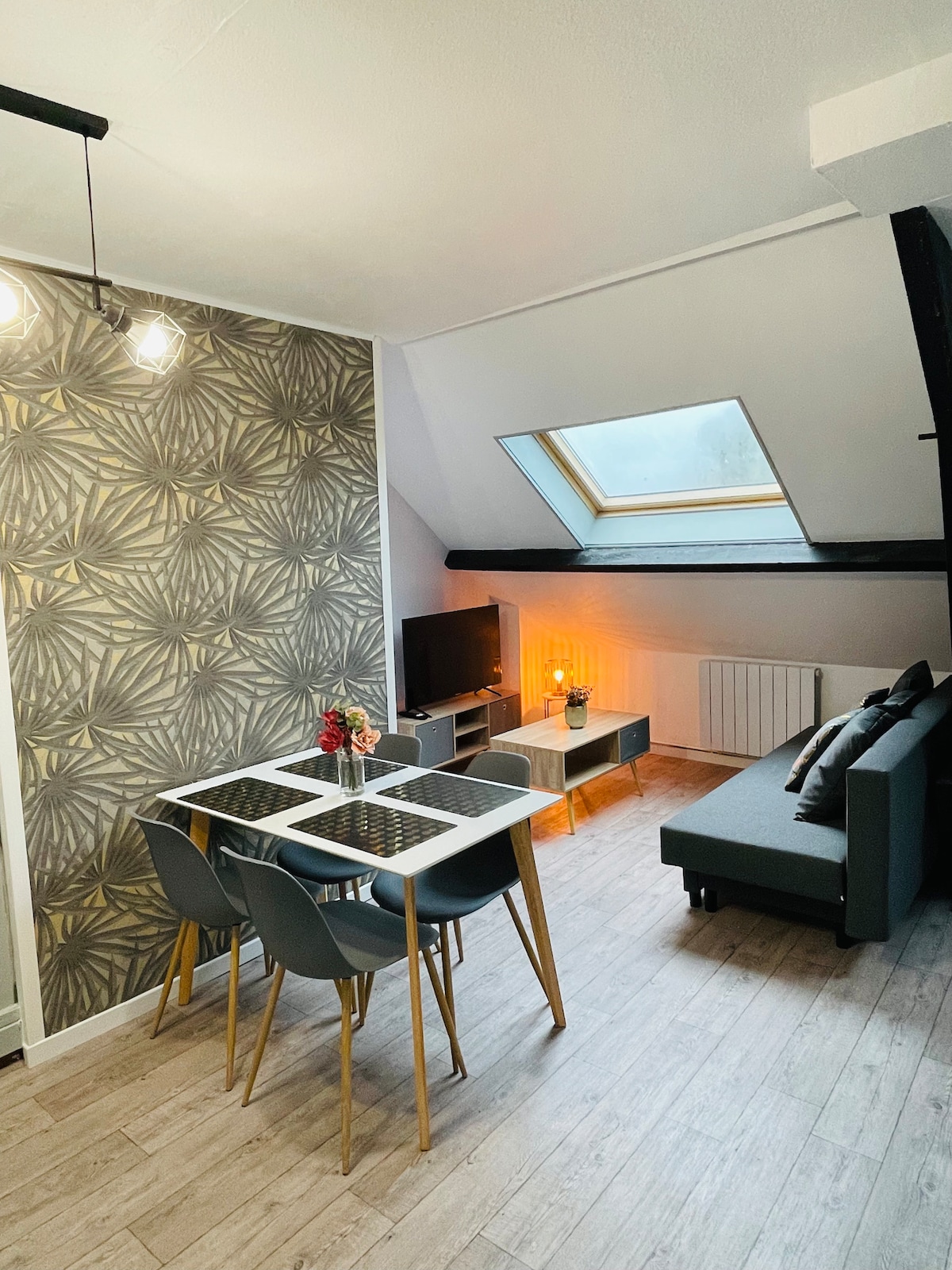 A dining area features a glass table surrounded by four chairs, positioned near a cozy seating area with a dark sofa. A modern television is mounted on the opposite wall, with warm light emanating from a side table lamp. Natural light filters through a skylight above.