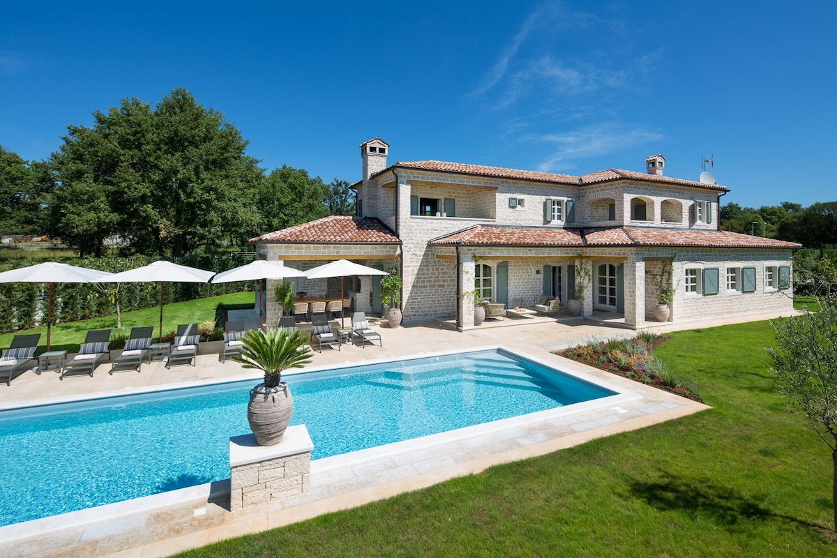 The villa's exterior features a large swimming pool surrounded by sun loungers and shaded by umbrellas. A landscaped garden provides a serene setting, with mature trees in the background. The stone structure displays a traditional architectural style with a tiled roof and multiple windows.
