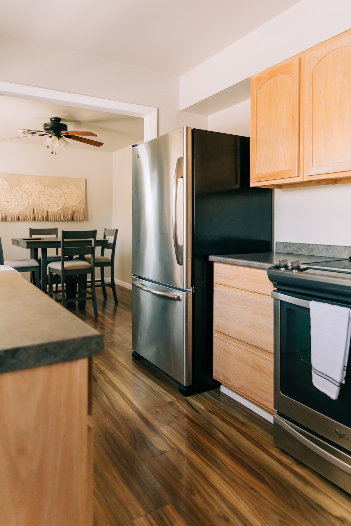 A modern kitchen area is visible, featuring stainless steel appliances including a refrigerator and stove. Light wood cabinetry complements the countertop. In the background, a dining table with four chairs is shown, alongside a wall adorned with textured art.