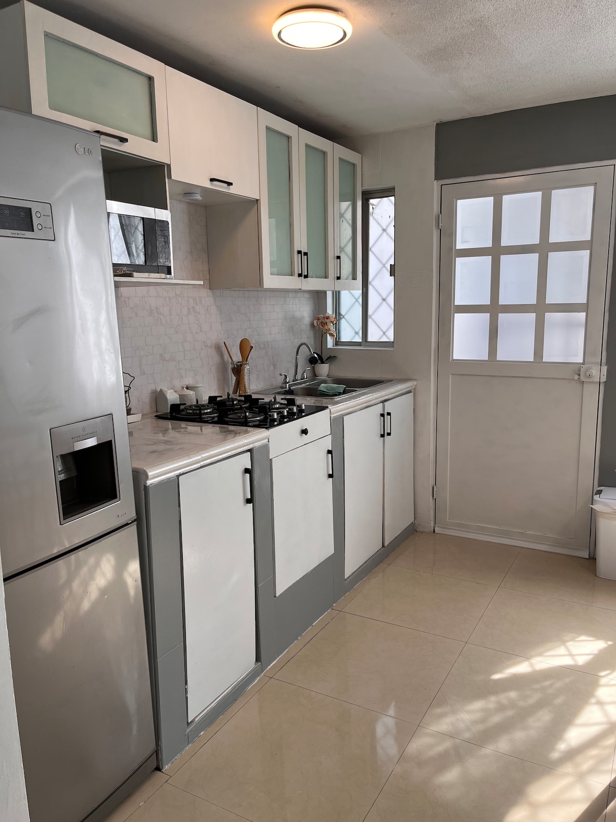 A modern kitchen is shown, featuring a stainless steel refrigerator and a countertop stove. Cabinets with glass fronts provide storage space, and a small window allows natural light to fill the room. The floor is finished with glossy tiles, enhancing the area’s brightness.