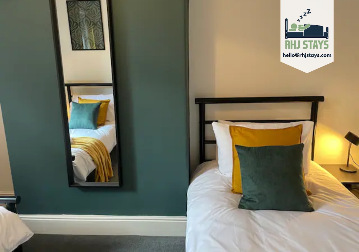 A cozy bedroom features a single bed with crisp white linens and a decorative yellow and green throw pillow. A large mirror on the wall reflects the room's calming green accent wall, enhancing the sense of space and light.