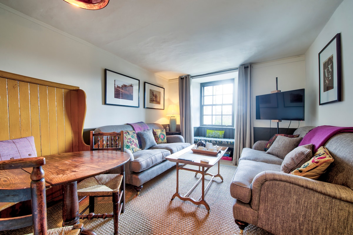 The living area features two comfortable sofas adorned with decorative cushions, alongside a wooden table surrounded by chairs. Natural light enters through a large window, which provides a view of the outside. Wall art enhances the space while creating a welcoming ambiance.