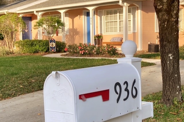 A white mailbox with the number 936 is prominently displayed in the foreground. Behind it, a charming cottage features light peach-colored siding and blue accents, including a front door. Neatly landscaped gardens with flowers and shrubbery are visible, creating an inviting entryway.