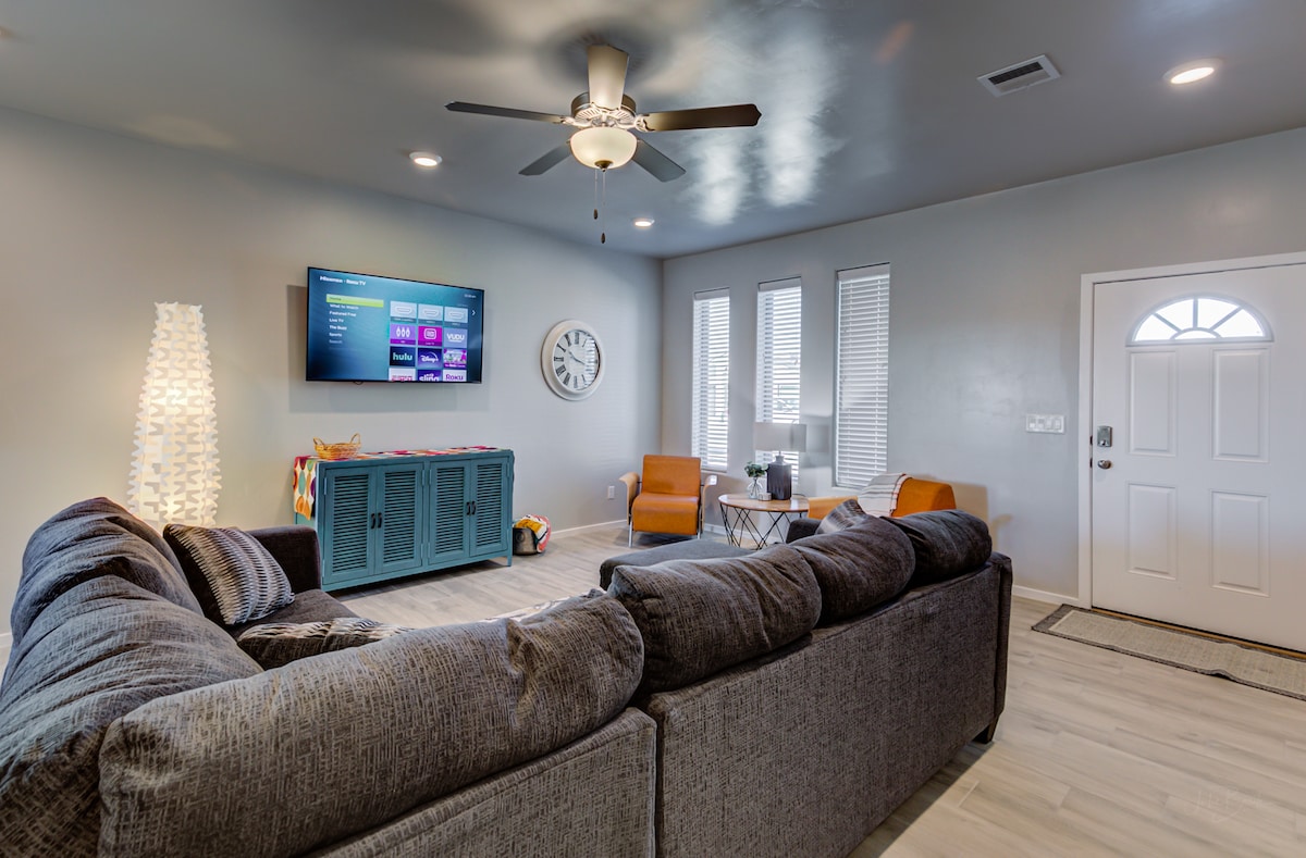 A spacious living room features a large gray sectional sofa and a flat-screen TV mounted on the wall. Natural light streams through multiple windows, and a decorative floor lamp adds soft illumination. A small table with two orange chairs is positioned near the entrance.