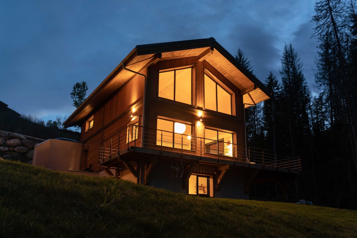 The chalet is situated on a grassy slope, illuminated against a twilight sky. Large windows allow warm light to radiate from the interior, while the wooden facade reflects a modern design. Surrounding trees enhance the natural setting.