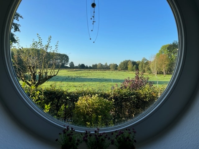 A circular window frames a serene view of lush green fields and trees under a clear blue sky. The window's edge is adorned with greenery, adding a touch of nature to the tranquil landscape.