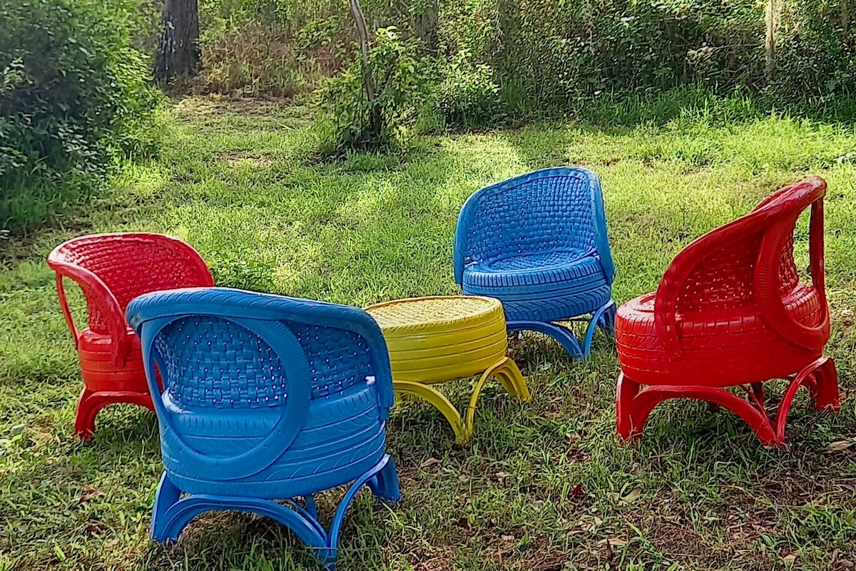 A circular arrangement of four colorful chairs is set on a grassy area. Two chairs are bright red and two are vibrant blue, surrounding a small round yellow table. Lush greenery and trees gently frame the peaceful outdoor setting.