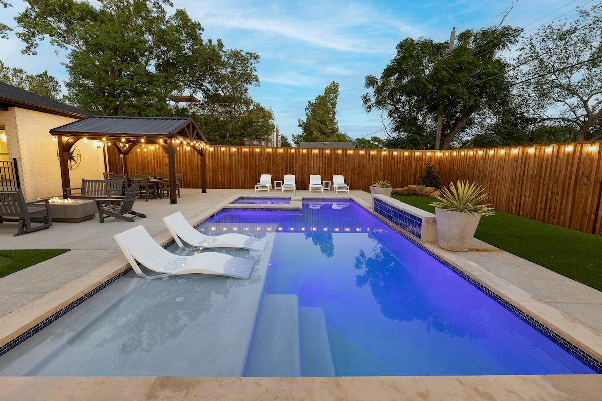 A spacious backyard features a resort-style swimming pool with a cascading spa. Lounge chairs are positioned around the pool, and soft lighting is strung along the perimeter. Lush greenery and a wooden fence provide privacy, creating a relaxing atmosphere for gatherings.